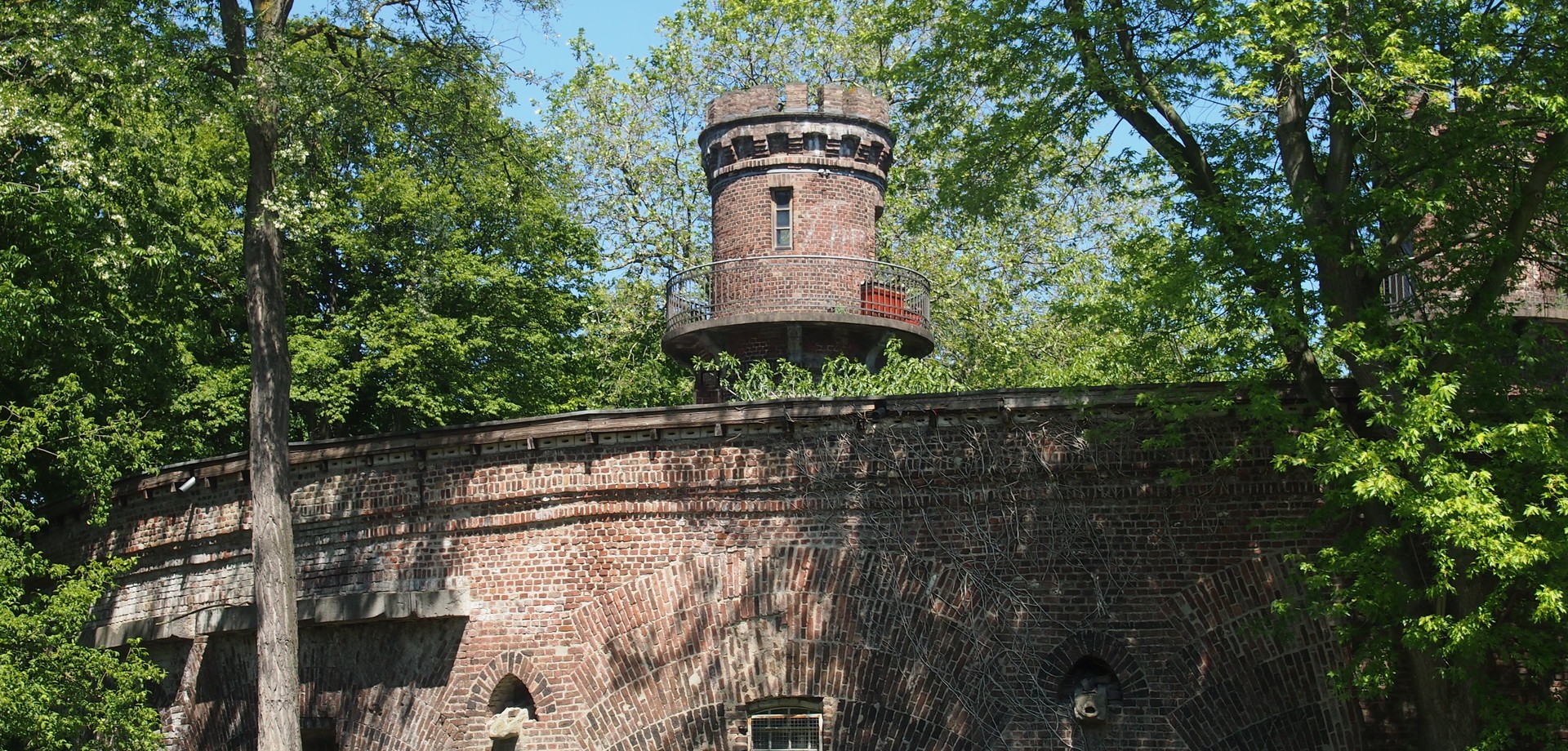 Eine alte Backsteinmauer im Grünen hinter der ein Turm aus Backstein hervorragt.