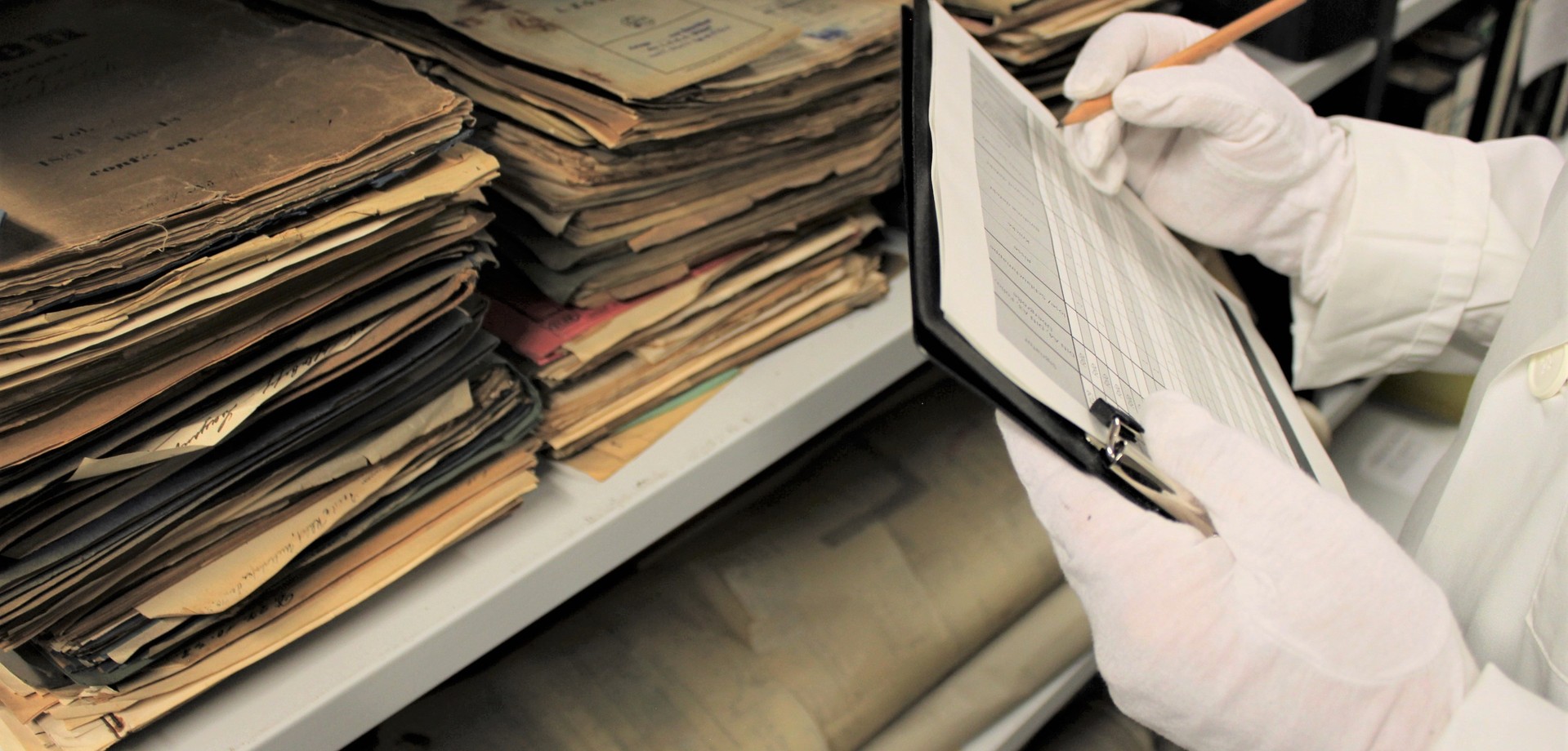 Alte Akten und Dokumente in einem Regal. Davor sieht man ein Klemmbrett, das von einer Person mit weißen Spezialhandschuhen zur Arbeit mit alten Dokumenten festgehalten wird.