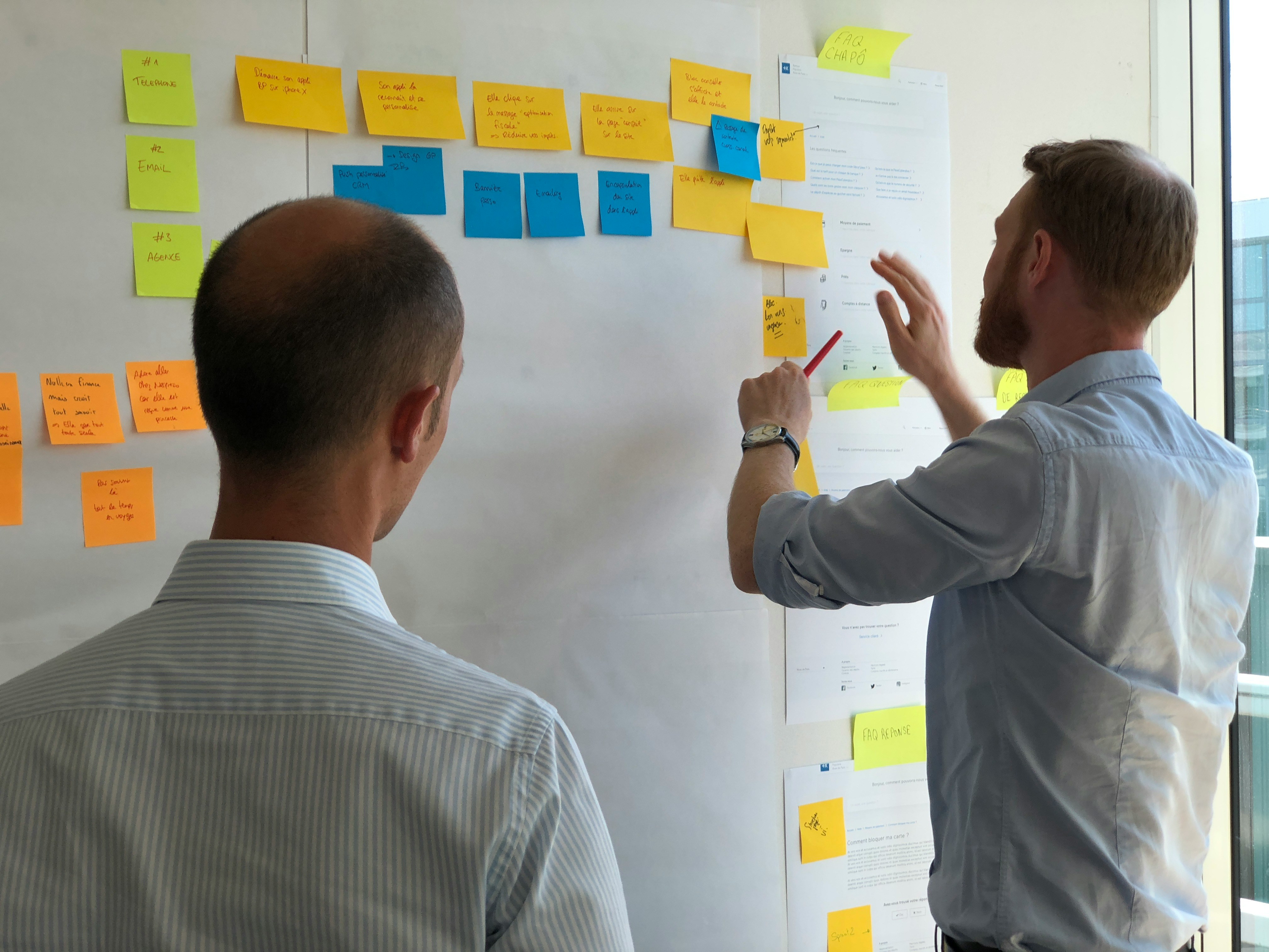 Zwei Personen stehen vor einem Whiteboard; eine Person klebt Post-Its auf das Whiteboard