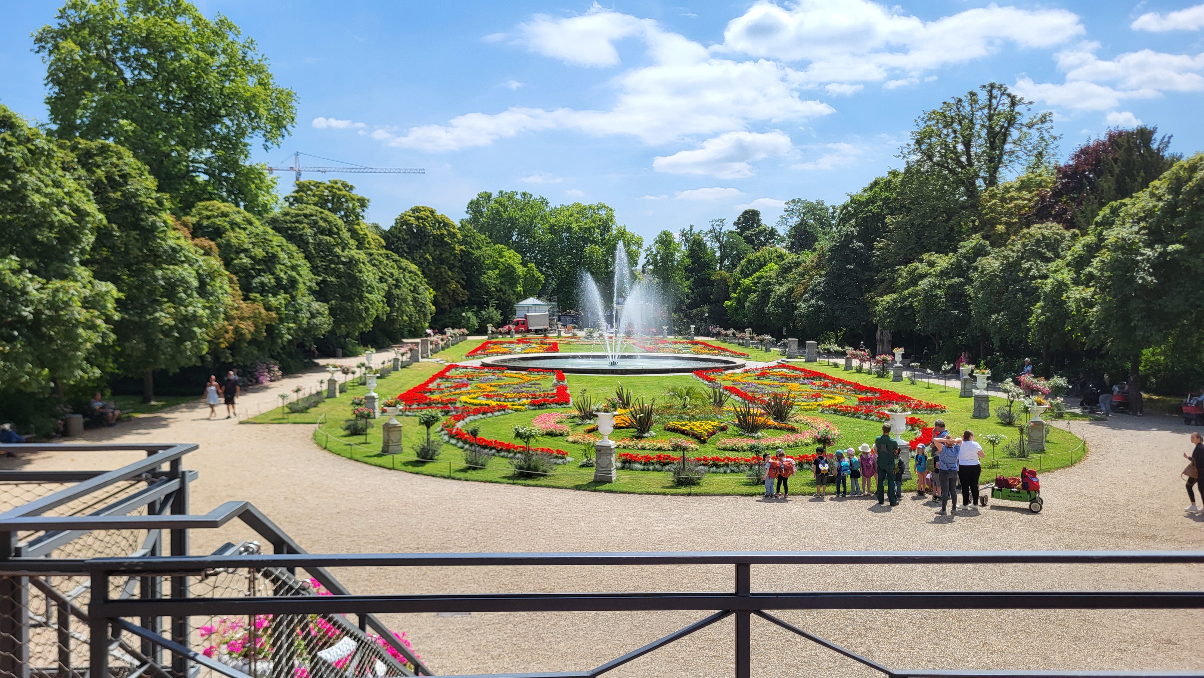 Ein großer Springbrunnen inmitten einer Grünanlage mit Gras und bunten Blumen. Drumherum ein breiter Kiesweg für Fußgänger und Bäume.