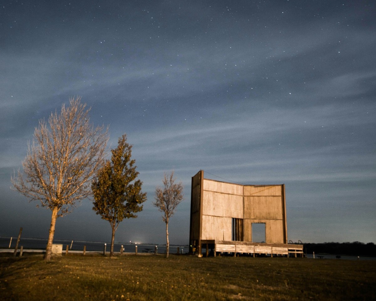 Eine Bühne aus Holz steht auf einem Feld bei Nacht. Daneben stehen drei unterschiedliche Bäume in einer Reihe.