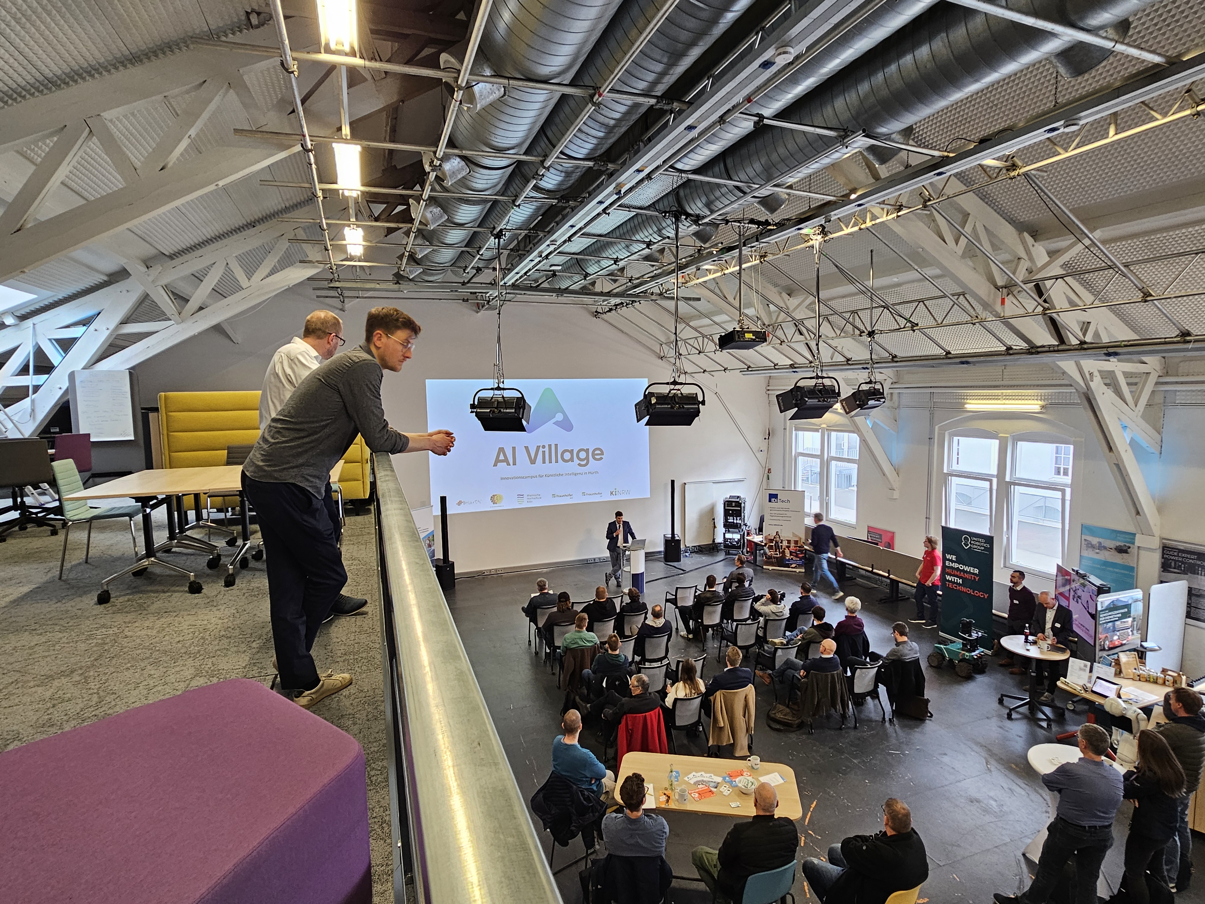 In einer Halle sitzen viele Menschen und hören einem Vortrag zu. Links oben auf einer Empore stehen zwei Männer und gucken auf die Menschen herab.