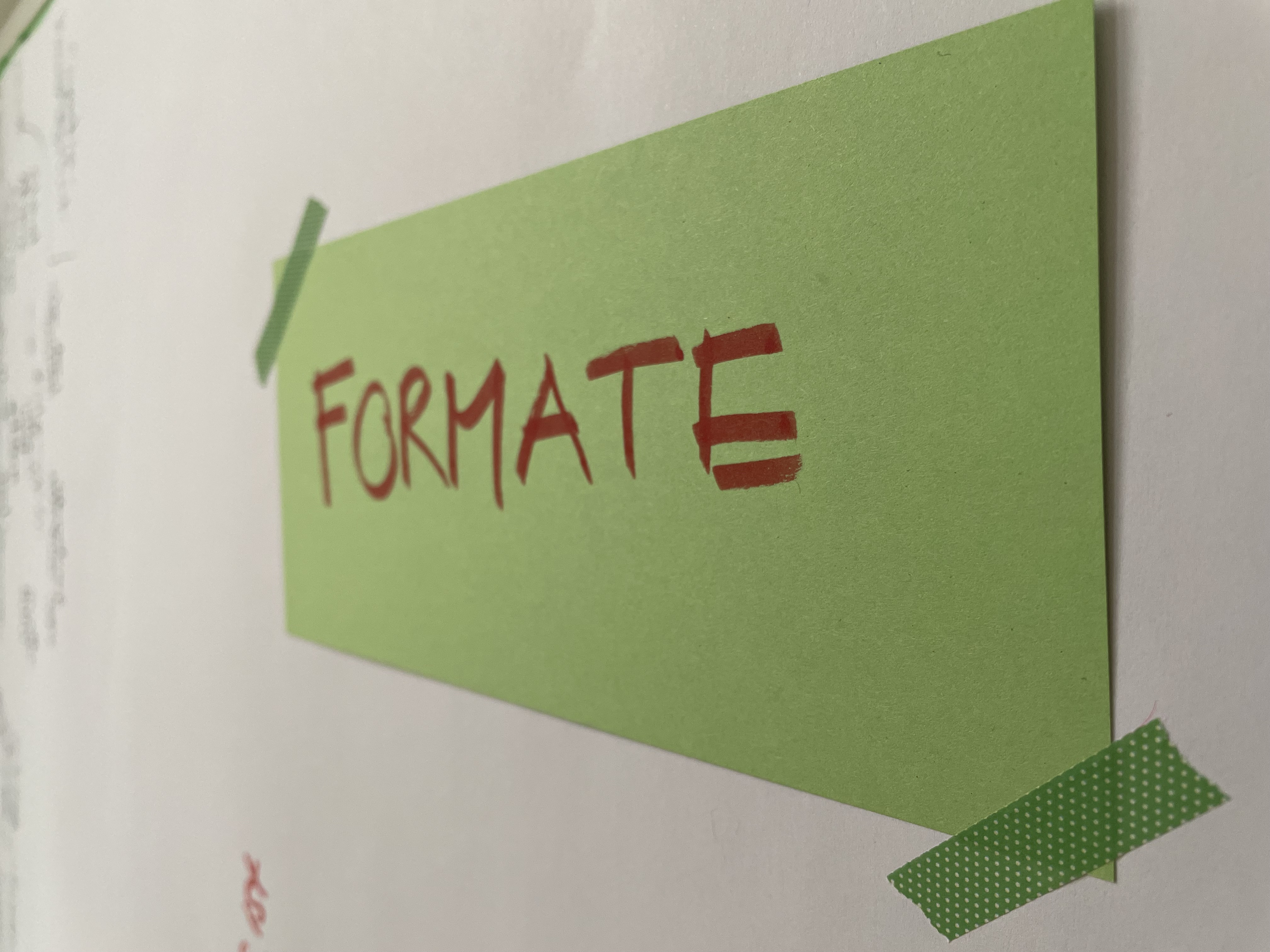 Ein grüner, rechteckiger Zettel klebt an einer Wand. Darauf ist mit rotem Stift das Wort "Formate" geschrieben