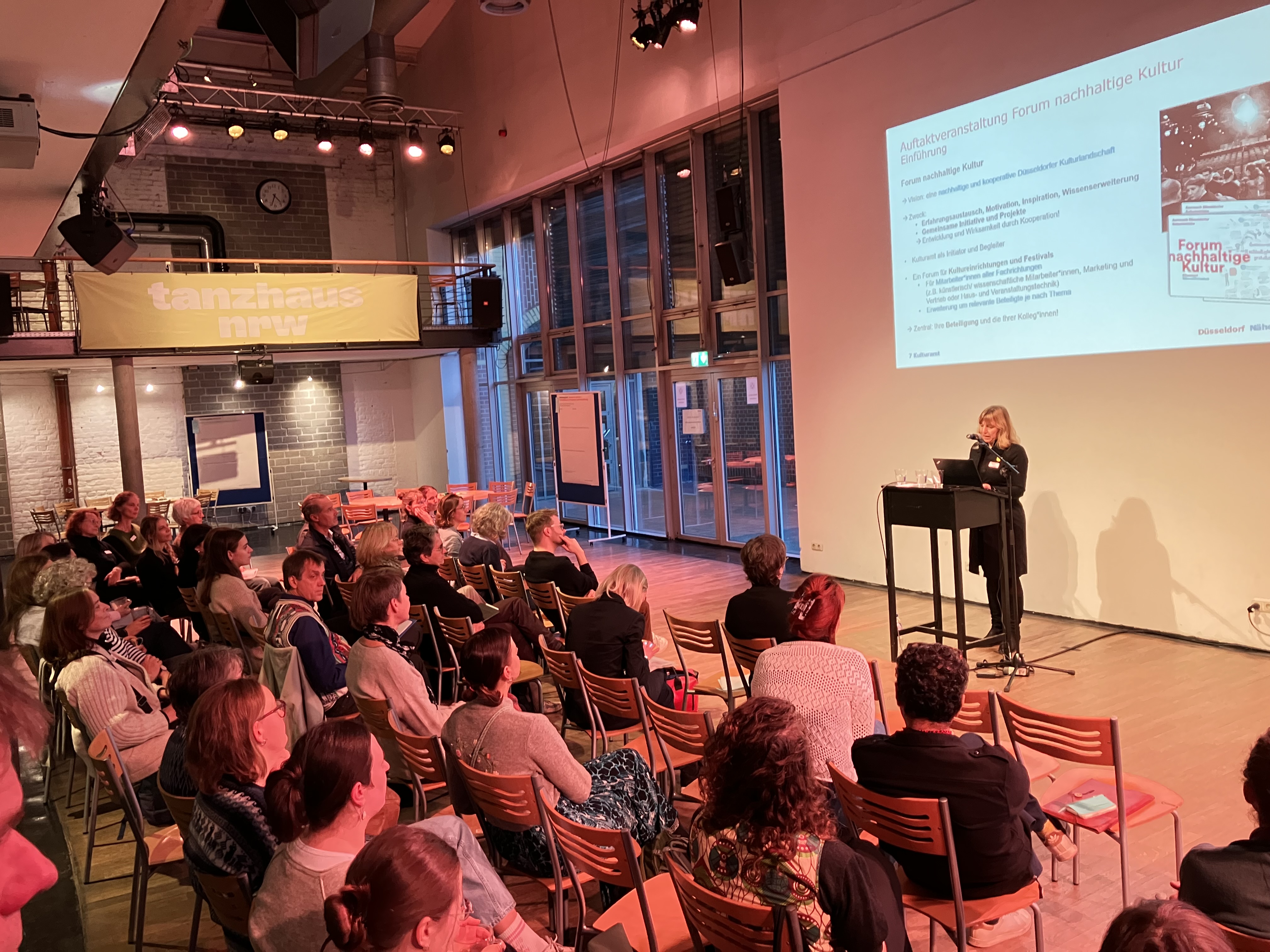 Viele Menschen sitzen im Publikum und schauen auf eine Bühne mit Rednerin.