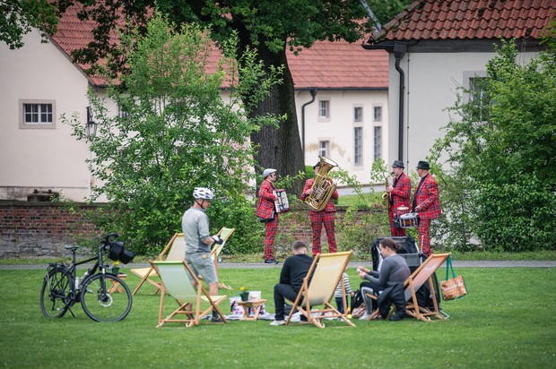Vier Personen in rot-kartierten Anzügen machen Musik im Grünen vor einer Gruppe von Menschen auf Liegestühlen