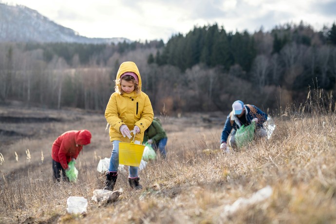 Erwachsene Personen und ein Kind in einem gelben Regenmantel sammeln Müll von einem Feld auf.