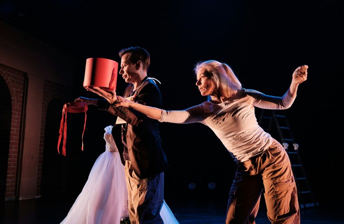 Zwei Personen in einer Theaterszene auf dunkler Bühne mit roten Requisiten, weißem Kleid im Hintergrund und dynamischer Körperhaltung
