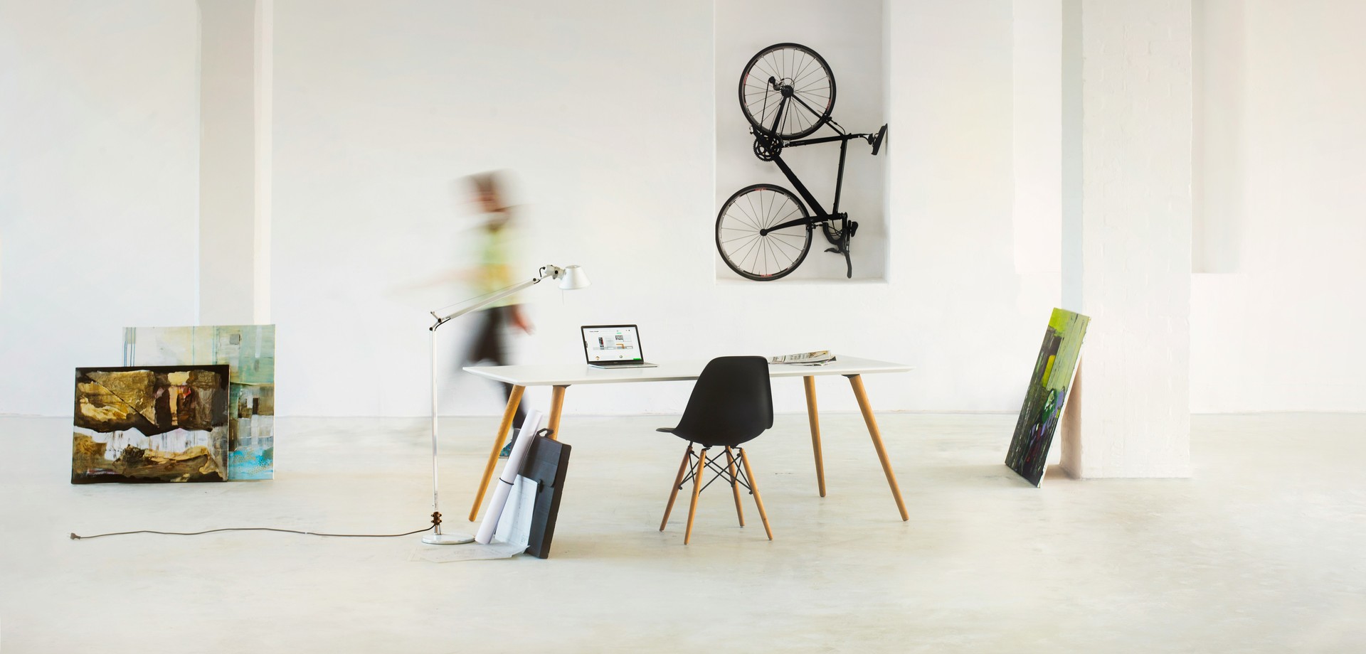 Arbeitsplatz  in einem hellen Raum mit Tisch, Stuhl und Laptop und einer verschwommenen Person, die dahinter läuft. Zudem sind Bilder und ein Fahrrad an die Wände gelehnt.