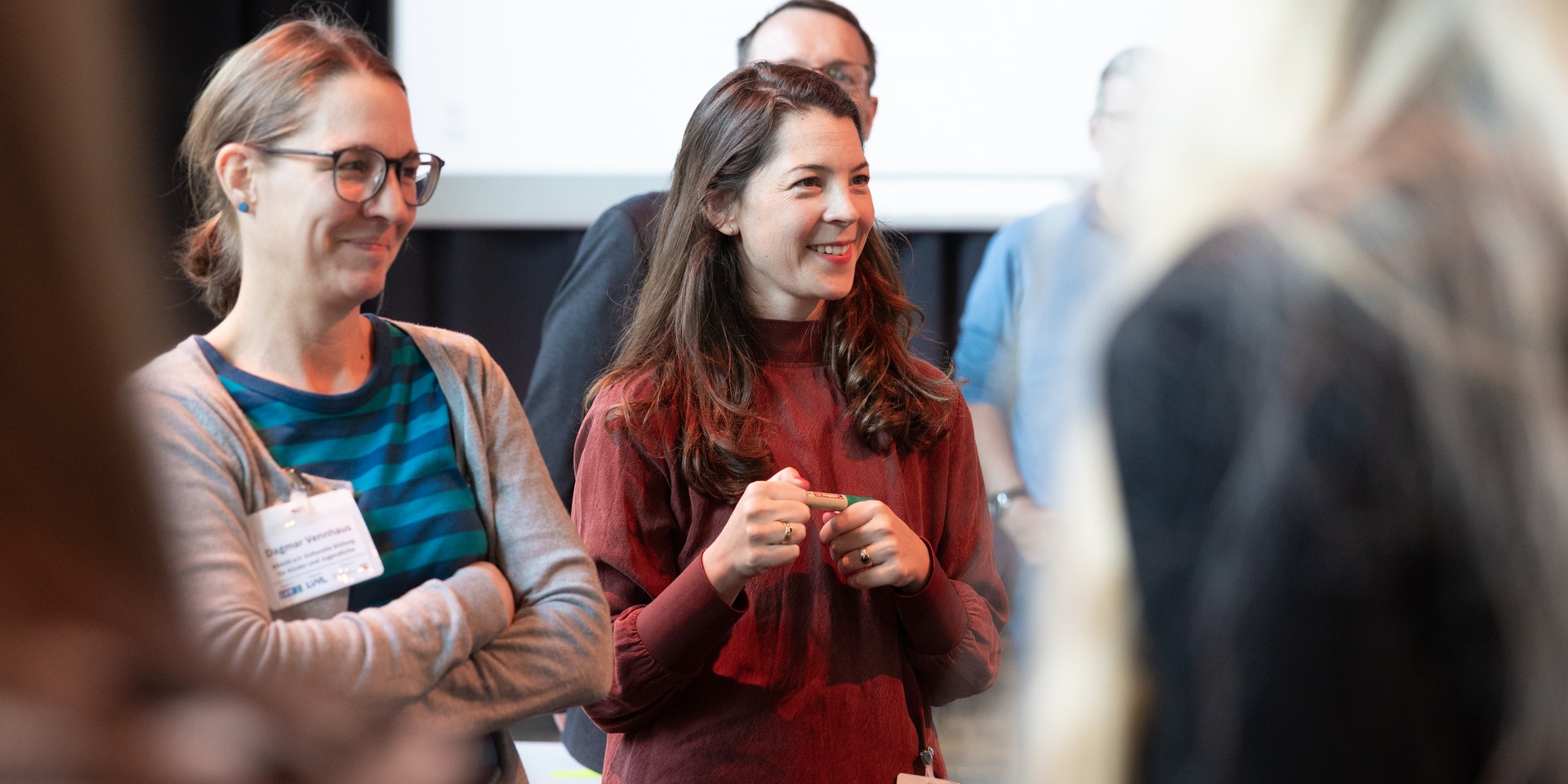 Frau steht und spricht mit einem Stift in der Hand umgeben von weiteren Personen