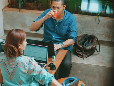 Zwei Personen sitzen an einem Tisch. Die Frau zeigt nach rechts und der Mann trinkt aus einer orangenen Tasse.