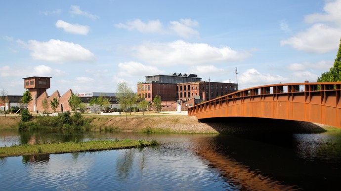 Metallbrücke führt über Fluss, im Hintergrund Backsteingebäude
