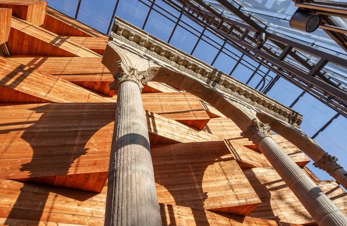 Blick nach oben an einer Holzfassade mit hervorstehenden Elementen entlang. Vor der Holzfassade stehen Überreste eines römischen Torbogens.