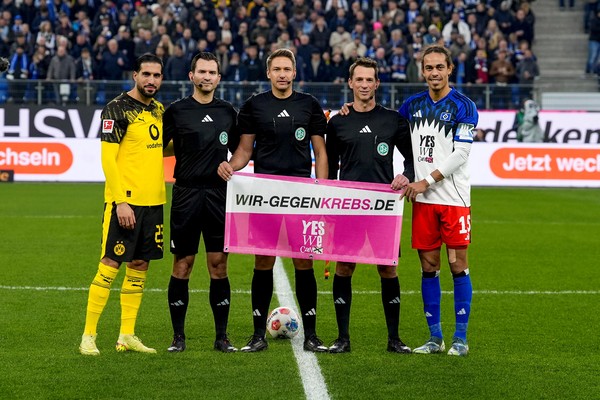 Zwei Fußballspieler und drei Schiedsrichter stehen auf dem Spielfeld und halten ein Banner der Initiative „wir-gegen-krebs.de“