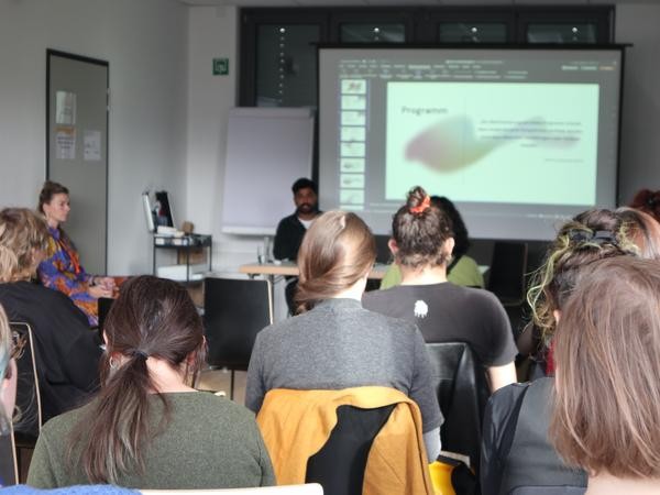 Blick von hinten auf eine sitzende Gruppe von Menschen, die nach vorne auf den Referenten und seine Präsentation schauen.