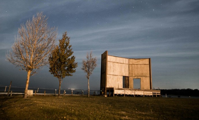 Eine Bühne aus Holz steht auf einem Feld bei Nacht. Daneben stehen drei unterschiedliche Bäume in einer Reihe.