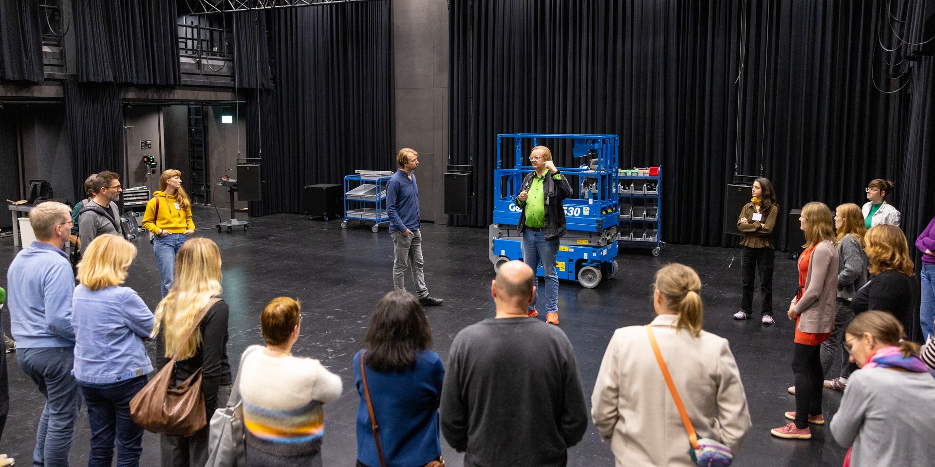 Eine Gruppe von Personen steht auf einer Theaterbühne mit einer blauen Hebeanlage
