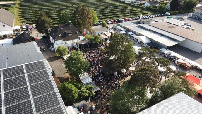 Luftaufnahme eines weitläufigen Open-Air-Festivalgeländes mit Bühne, dichtem Publikum, Zelten, Gebäuden, Solaranlagen und umgebenden Grünflächen.