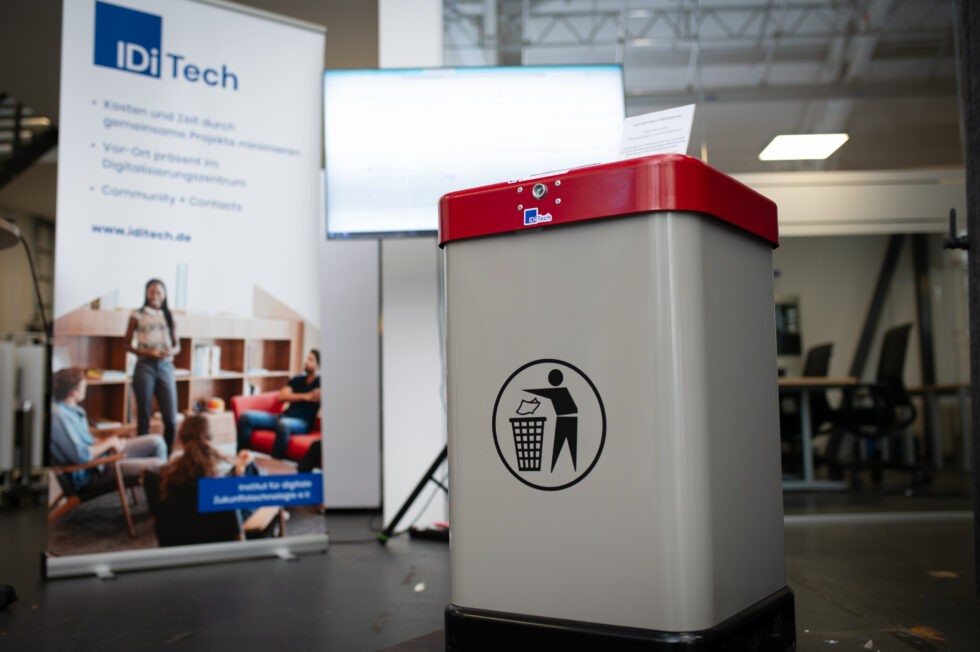 Im Vordergrund steht ein KI-gesteuerter Sensor-Mülleimer. Im Hintergrund sind ein Werbeaufsteller von IDiTech und ein Bildschirm zu sehen.