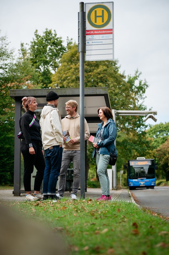 Vier Personen warten an einer Haltestelle auf den Bus.