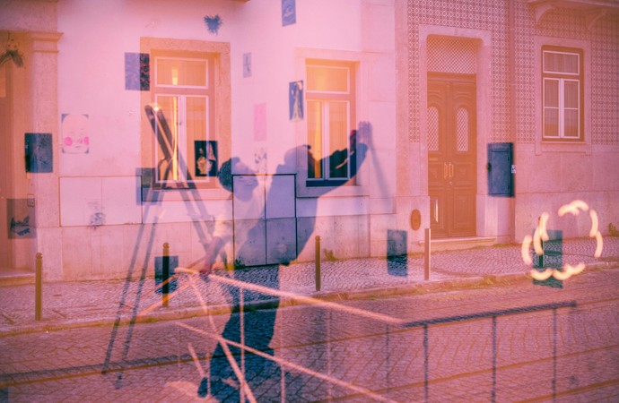 Rosa eingefärbte Spiegelung einer Straßenszene mit Hausfassade, Fenstern, Eingangstür und schemenhaften Personenreflexionen im Vordergrund