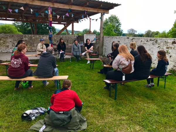 Eine Gruppe von Menschen sitzt bei einem moderierten Gespräch im Freien auf Bänken und im Gras.