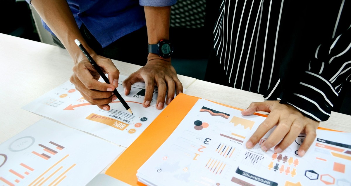 Zettel mit Daten liegen auf einem Tisch, die Hände von vornübergebeugten Menschen sind zu sehen.