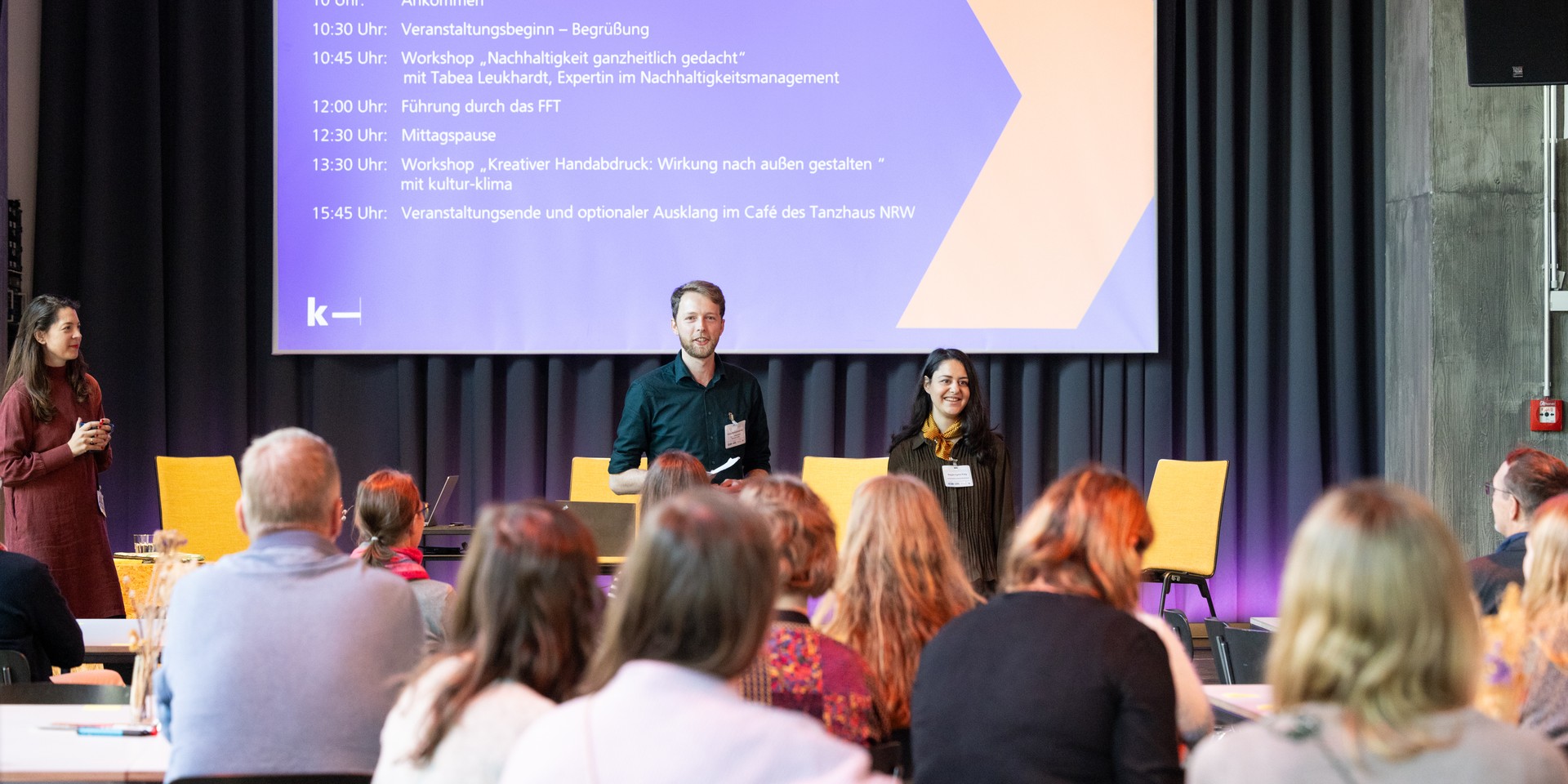 kultur-klima Team aus zwei Personen steht und spricht vor Teilnehmer:innen, die in Tischgruppen zusammensitzen.
