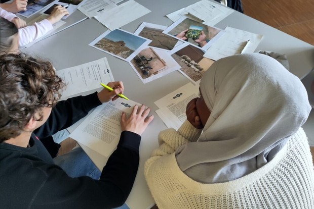 Ein Jugendlicher mit kurzen braunen Haaren und eine Jugendliche mit Kopftuch sitzen an einem Tisch mit Fotos und Schriftdokumenten.