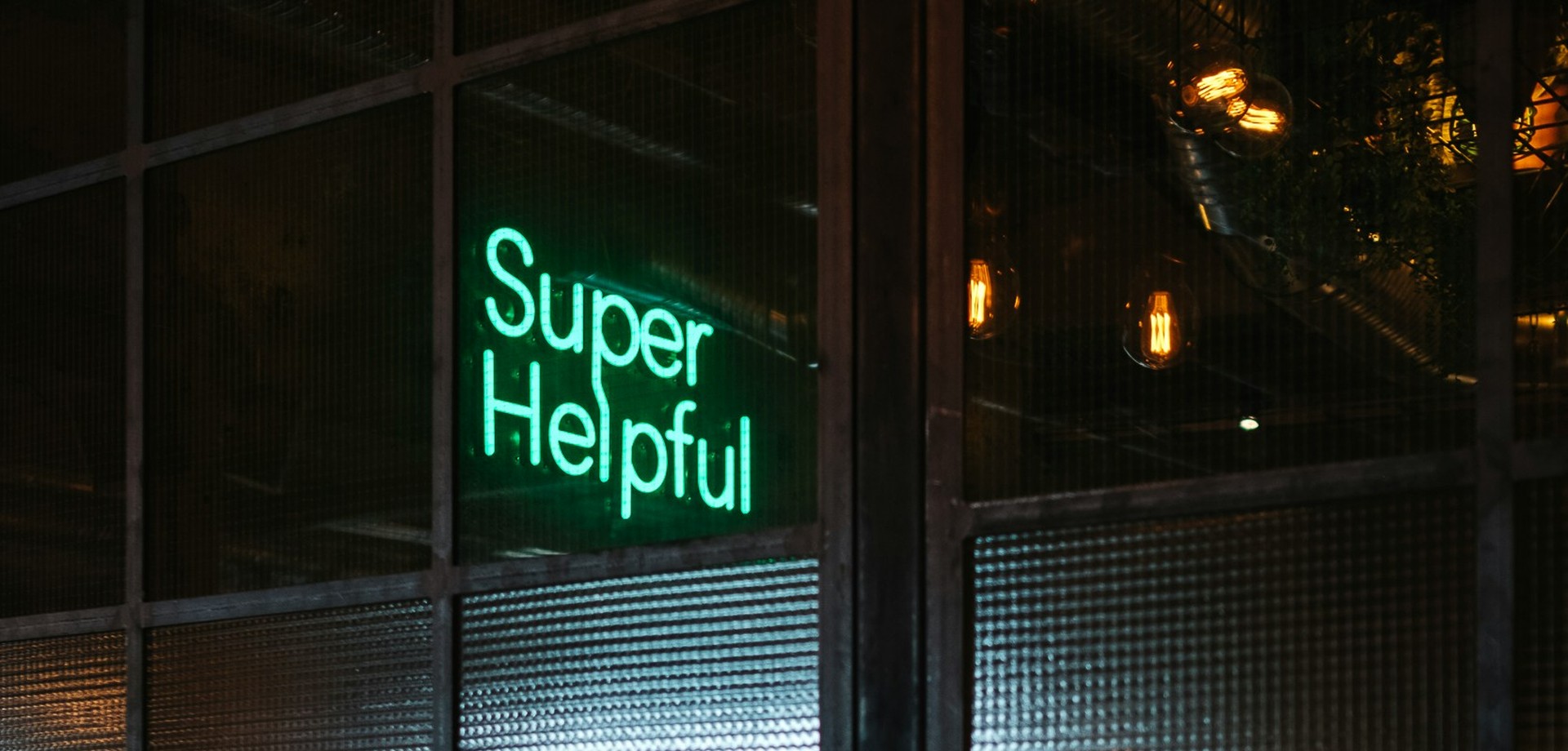 Dunkles Sprossenfenster, durch dieses sieht man einen türkisen Neon-Schriftzug: "Super Helpful"
