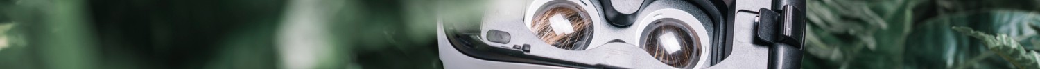 Lächelndes blondes kleines Mädchen mit VR-Brille steht unter einer Grünpflanze.