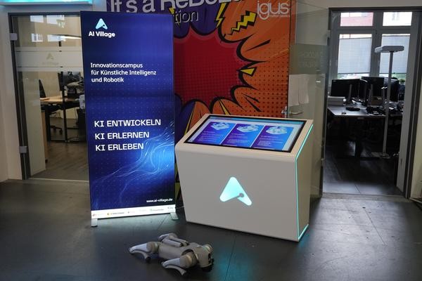 Ein Roboterhund liegt vor einer Infotafel (rechts) sowie einem Werbeaufsteller (links) von AI Village.