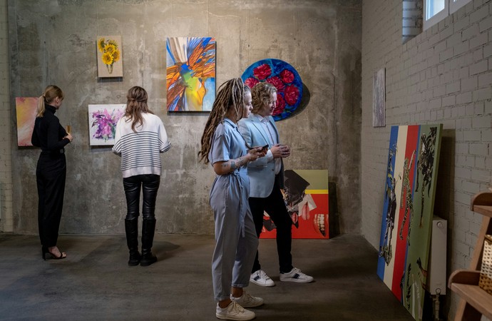 Personen betrachten Kunstwerke in einem Ausstellungsraum; an den Wänden und am Boden sind verschiedene farbige Gemälde zu sehe