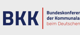 Das Logo der Bundeskonferenz für Kommunalarchive. Es besteht aus dunkelblauen Buchstaben auf hellgrauem Grund mit einem rötlichen Zierstrich.